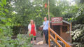Die Kita-Leiterin Christiane Klar und Corinna Rüffer im Garten der Kita umgeben von Bäumen und Büschen. An einem Pfahl neben ihnen ist ein Holzschild befestigt, auf dem steht: „Sinnespfad“.