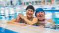 Eine ältere Frau und ein älterer Mann, Arm in Arm am Beckenrand eines Schwimmbeckens in einem Hallenbad.