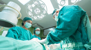 Das Bild zeigt einen OP-Raum/-Tisch in einem Krankenhaus. Drei Personen in OP-Kleidung beugen sich über einen OP-Tisch, auf dem eine Person liegt,.