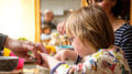 Das Bild zeigt ein Kind, dass mit seinen Eltern am Esstisch sitzt. Die Mutter sitzt neben dem Kind. Vom Vater ist nur die Hand zu sehen, mit der er dem Kind beim Essen hilft.