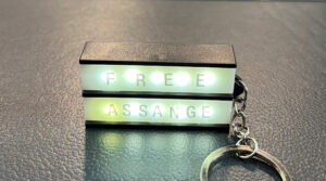 Auf einem Tisch liegt ein Schlüsselanhänger, auf dem in Leuchtschrift steht: FREE ASSANGE.
